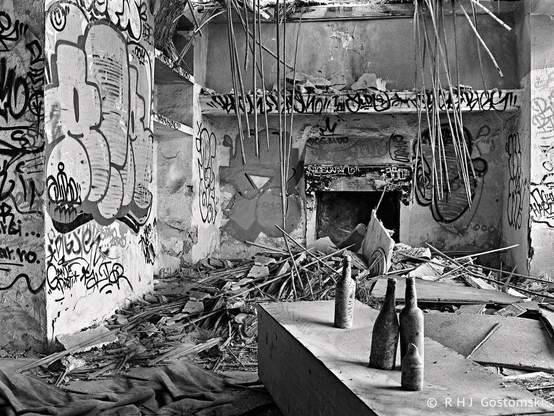 A black and white photo inside an abandoned farmhouse. The ceiling has collapsed and there is graffiti on the walls. Three dusty beer bottles stand on an overturned cupboard in the foreground. 