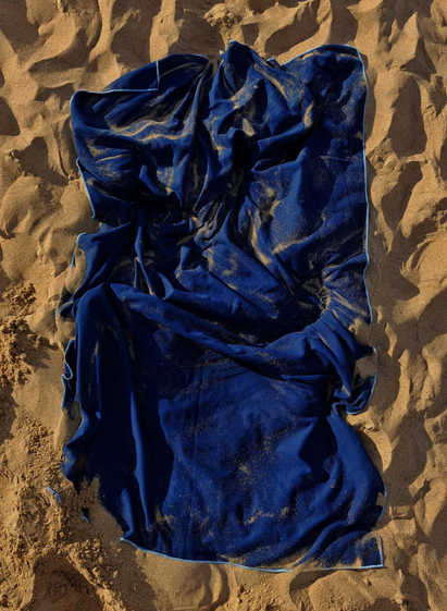 A crumpled blue fleece towel lies on sandy ground, covered with sand particles. The towel is  full of dents and folds with darker shadows and it's blue cloth contrasts against the light brown sand surface.