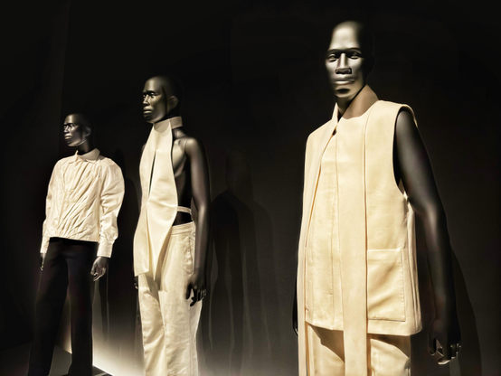 A display of three identical, dark-toned male-presenting mannequins wearing different designer outfits in white or cream hues against a black curtained backdrop. Part of the Metropolitan Museum’s exhibition “Superfine: Tailoring Black Style,” about the history and current expressions of Black dandyism. Designer info from left to right, from the museum placards:

1. Bianca Saunders
Ensemble, autumn/ winter 2024-25
Shirt of white cotton plain weave, trousers of black wool-silk twill

2. Maximilian Davis 
Ensemble, spring/summer 2021
Top of white cotton poplin, trouser of white cotton drill

3. Ib Kamara 
Ensemble, spring/summer 2023
Sleeveless jacket and trousers of cream wool twill
