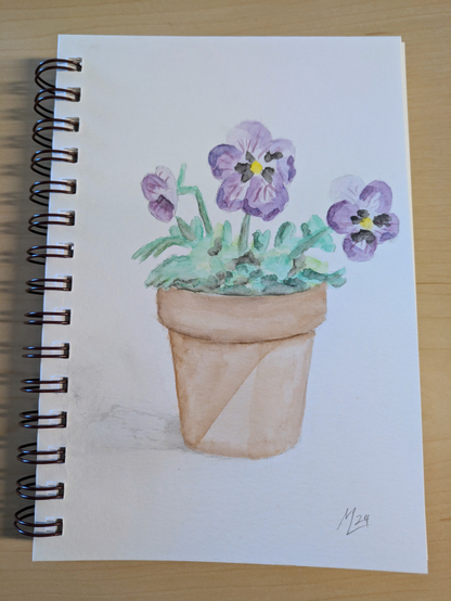 This is a watercolor of a pansy in a pot. It's done in a notebook which you can see lying on a desk surface.