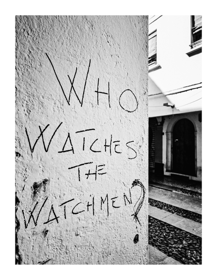 This is a black and white  photograph which features a textured concrete wall with graffiti reading "WHO WATCHES THE WATCHMEN?" in bold, handwritten letters. In the background there is a cobblestone street and in the other side of it there is an arched doorway of a building. capturing a European urban setting.