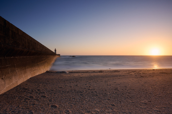 Upfront a sandy beach, then a milky sea and some rocks within the sea. On the left side a wall is leading into the ocean with a lighthouse on the end. The horizon is in the middle and on the right the  setting sun.