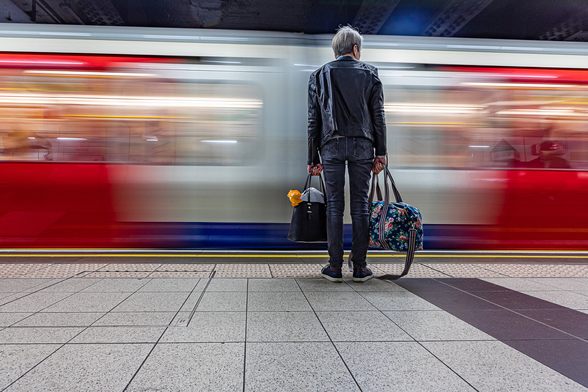 A man dressed in black is waiting on a metro platform for a blurry blue white and red metro train to stop in front of him, in a London tube station. His holding two hand luggages. The train is blurry due to the use of a slow shutter speed.