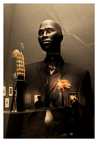 A scene from the Metropolitan Museum’s exhibition “Superfine: Tailoring Black Style,” about the history and current expressions of Black dandyism. Closeup on a bald mannequin, from torso up, in a dark pinstriped jacket with a posy of colorful flowers pinned to the lapel (1). The display’s protective glass reflects an identical mannequin across the room, wearing a black coat with gold braid and fringed epaulettes over tracksuit pants (2). A third mannequin is somewhat visible reflected against the jacket of the first one. The reflection also shows a person perusing framed artworks on the far wall. The museum lighting dramatically picks out elements of color against mostly subdued darker tones.

Footnotes (info from the museum placards):
(1) Emerie Tchatchoua in collaboration with Olga Prinku, “Suit,” 2021: Jacket of navy pinstriped wool gabardine embroidered with bouquet of polychrome dried flowers; trousers of navy pinstriped wool gabardine

(2) Olivier Rousteing, “Ensemble,” 2023: Coat of black cotton velvet with fringed epaulets and Brandenburg closures of gold metallic braid with embroidered pearls and crystals; tracksuit of black, white, and gold polyester satin