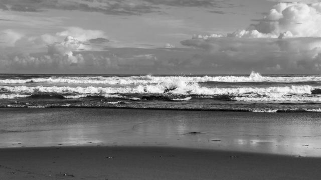 Black and white view of a beach with waves crashing under a partly cloudy sky. Wet sand reflects the horizon and clouds.