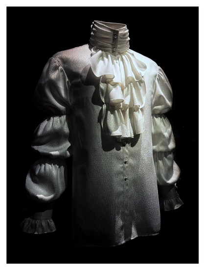 A view of the entire shirt against a black backdrop. The dramatic lighting again highlights the layered ruffles of the jabot and the puffed sections of the sleeves.

From the museum placard: “During the period of his 1984 album and film Purple Rain, ruffled Regency-style shirts and closely cut tailcoats became his signature — a New Romantic echo of early nineteenth-century dandyism and the peacock styles of 1960s London. This top, designed by Marie France, made by Louis Wells and Vaughn Terry Jelks, and worn by Prince during the Purple Rain tour, features pearl buttons and bejeweled ‘cuff links,’ touches that enhance the garment's exuberant dandyism.”