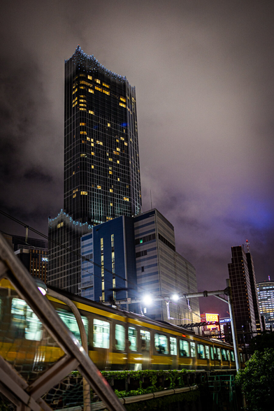 A train illuminated from inside is speeding at night in front of a skyscrapper under a cloudy sky creating a cyber punk mood, in Shinjuku, Tokyo, Japan.