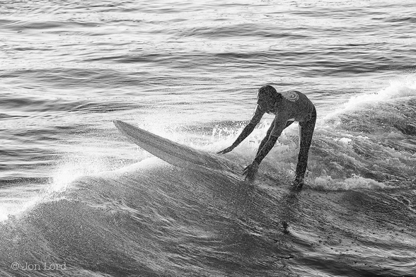 This is a black and white photo of a young woman surfing shortly after dawn.
To the right of centre is a young female surfer standing on her surfboard with feet astride and bending forwards with her hands holding the edges of the board. She is looking ahead, moving from the right to the left. She is wearing a t-shirt and wetsuit trousers, perhaps the T-shirt is covering a wetsuit top? The board is white with two near parallel black stripes running along the length. The girl is partially obscured by fine water spray. Our surfer is on the crest of a breaking wave that stretches diagonally across the image from the lower left corner towards the upper right. The water behind the wave is disturbed while the water ahead is smooth.
The location: Hermosa Beach, California (2013).