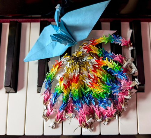 Auf einer Klaviertastatur liegt ein zu einem Bündel verbundenes und in Regenbogenfarben kombiniertes Origamiwerk aus dutzenden Mini-Kranichen.
Die Normalgröße darüber in hellblau. 