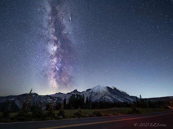 A breathtaking night sky displays the Milky Way stretching across the stars above Mount Rainier / Tahoma in Washington state with an iridium flare.
Snow-capped peaks and pine trees stand silhouetted against the horizon, with a road leading into the scene at the Sunrise corridor of the Mount Rainier National Park.
A climber is already on the way to the peak, indicated by their headlights on the side of the mountain at 12:23AM on July 29, 2025.