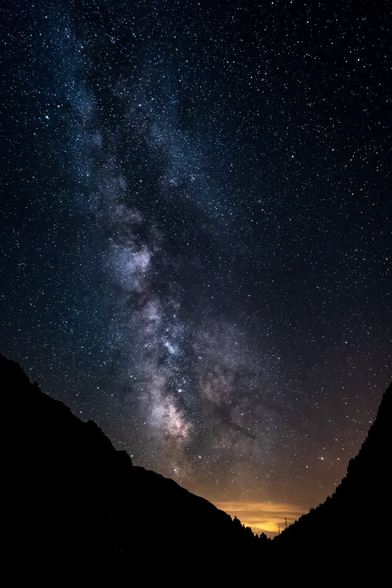 Starry night sky with the Milky Way visible in the center, framed by dark mountain silhouettes. An orange glow on the horizon.