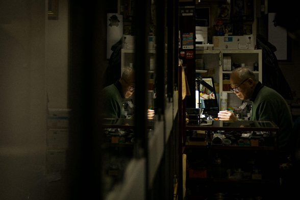 Inside a dark camera shop, a desk light spotlights a Japanese man working meticulously on a vintage camera. A reflection of him is displayed on the cabinet glass doors in front of him.