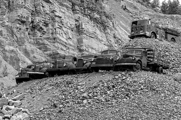 A black and white landscape photo of the front ends of six 1950s era pickup trucks parked tightly together on a small hill of rock rubble in a stone quarry. Their front ends stick out slightly over the edge of the hill. They cut across the image from left center to right center. The trucks on the left are slightly behind the ones on the right. Above and to the right of the trucks is a 1970s era pumper style fire engine. It is parked on a second hill of stone quarry rubble and points in the same direction as the pickups, toward the left. The background is a vertical cliff face of rock.