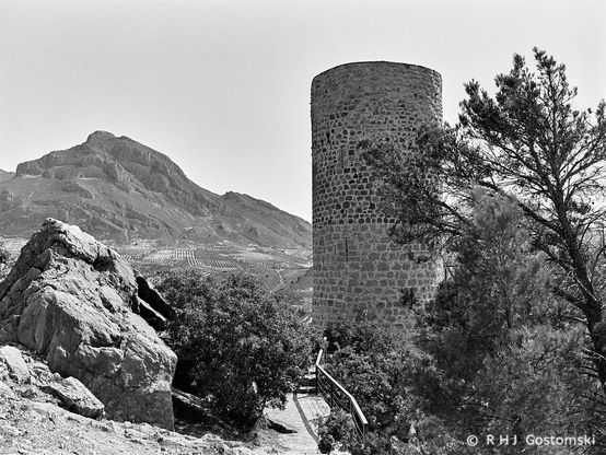A black and white photo of a path leading to a stone watch tower. A large rock outcrop is on the left of the photo, and a mountain is in the distance.