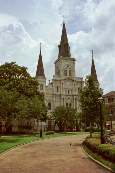 Colour negative film scan. A grand cathedral with three spires stands against a cloudy sky. A well-kept path and lush greenery lead to its entrance.