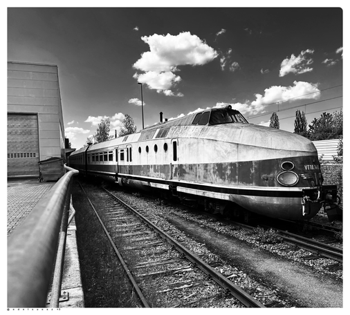 Schwarzweißfotografie: abgestellter dieselhydraulischer Schnellverkehrstriebzug der Deutschen Reichsbahn aus den 1960er Jahren, DDR, ausgeblichene abgeplatzte Zweifarblackierung, windschnittiger Triebwagenkopf mit eierförmigen Frontlampen, davor noch ein Gleis und ein Geländer, darüber weiße Wolken und dunkelgrauer Himmel 