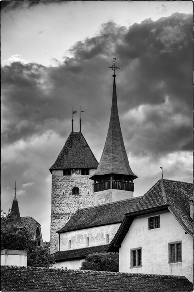 Schwarzweissfotografie im Hnochformat eines Kirchturms mit spitzem Dach und Wetterfahne neben einem steinernen Turm mit zwei Wetterfahnen, davor Häuser unter bewölktem Himmel