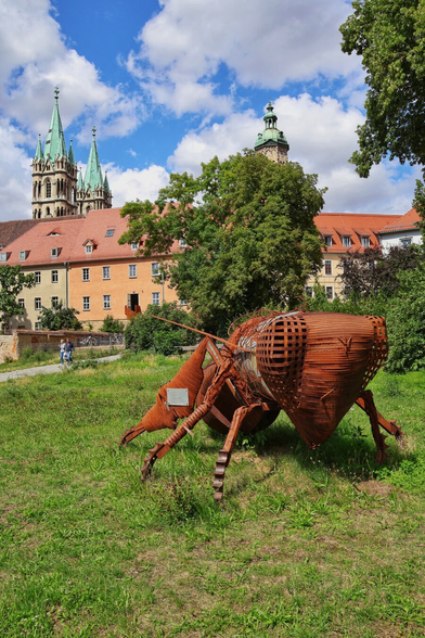 A striking sculpture of a large, rusty brown insect made of woven metal strips sits on a patch of green grass in the foreground. The insect is facing right, with its head and large, intricate eye visible. In the background, a large, multi-story building with orange-red walls and a red-tiled roof stretches across the middle of the frame. Two green spires of a cathedral rise above the building to the left, and a smaller, green dome is visible to the right. The sky is blue with scattered white clouds, and lush green trees partially obscure the buildings. A couple of people are walking on a path in the distance.