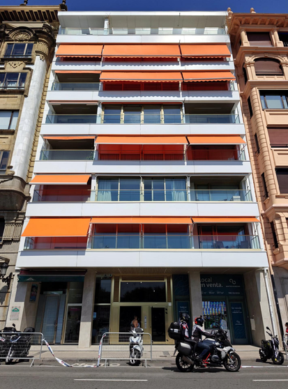 A modern white apartment building with orange and red retractable awnings on multiple balconies under a clear blue sky, flanked by historic buildings on either side. The ground level features storefronts. Motorcycles and scooters are parked on the street in front.