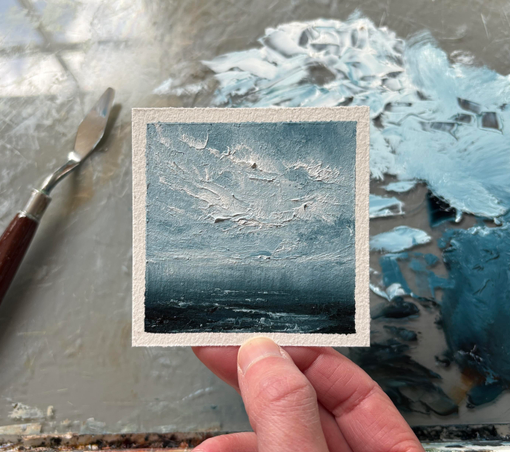 Photo of a hand holding an original tiny seascape oil painting by Tisha Mark, "Soothing Sea No. 2506" 3"x3" oil on Arches oil paper (2025). The painting is a tiny textured seascape with white atmospheric clouds over a deep blue sea. A paint palette with puddles of oil paint and a palette knife are visible in the background.