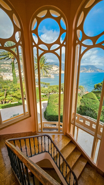 A staircase with ornate iron railings descends into a lush garden, with large, circular windows framing a breathtaking view of the Mediterranean coastline. The bright blue sky and distant mountains are visible through the windows, while palm trees and carefully manicured greenery surround the area. The walls are painted in soft pastel pink tones, and the floor is made of polished marble.
