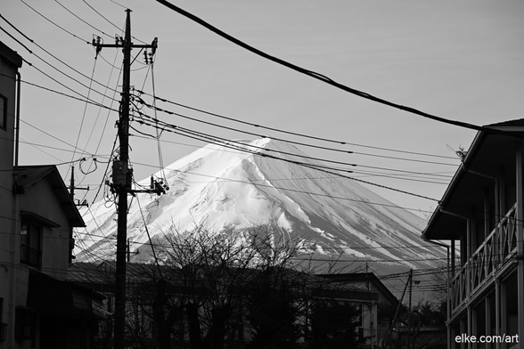 A striking black and white photograph of Japan's iconic Mount Fuji as seen from the town of Fujikawaguchiko.

The majestic snow-capped peak rises behind a foreground of silhouetted rooftops and a web of overhead power lines, creating a visually arresting contrast between nature's grandeur and urban infrastructure. The interplay of light and line draws the viewer’s eye toward the mountain, lending the scene both a sense of calm and quiet complexity.

This minimalist composition is ideal for modern interiors and lovers of fine-art urban landscape photography.

Perfect for living rooms, workspaces, or creative studios – especially those with a love for monochrome elegance and Japanese scenery.
