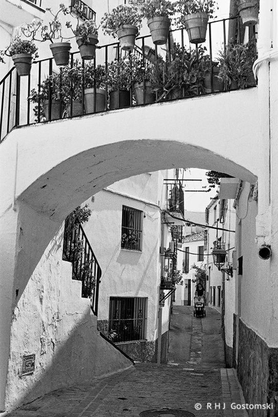 A black and white photo of a narrow cobbled street in Jubrique with white houses on both sides. In the foreground is an arch over the street, carrying a path to the front door of a house.
