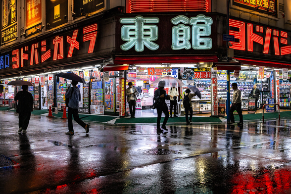 Dark silhouettes of people with umbrella going in different directions and backlit by a colourful shop covered in neon lights under a rainy evening in a Tokyo street. The scene is reflected in the wet pavement.
