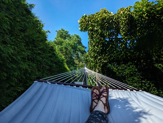 A first-person perspective shot from a white, quilted hammock, looking up at a clear blue sky. A person's feet, clad in dark red sandals with crisscrossing straps, rest on the end of the hammock. The hammock is strung between two lush green areas. To the left, a dense, dark green hedge forms a solid wall. To the right, a vibrant green vine climbs a wooden structure. The scene suggests a relaxed, sunny day in a garden.