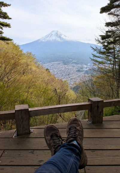 Im Hintergrund der an der Spitze mit Schnee bedeckte Mount Fuji. Der höchste Berg Japans.
Davor die Stadt Kawaguchiko in leichtem Dunst.
Eingerahmt von grün, gelber Vegetation, sitze ich auf einem Holzplateau in Jeans und braunen Turnschuhen. 