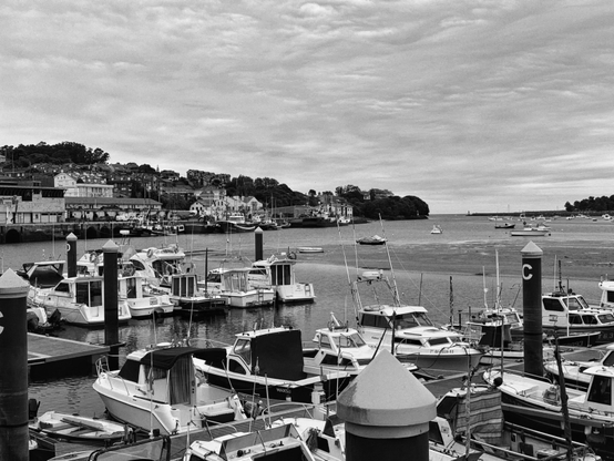 Una fotografía en blanco y negro de un puerto con numerosos barcos amarrados. En primer plano, se ven de cerca varios barcos de recreo y pesca, de tamaño pequeño a mediano, algunos de ellos atados a postes oscuros que tienen una letra "C" blanca. El agua del puerto parece tranquila, y en la parte media de la imagen, la marea baja ha dejado al descubierto áreas de lodo y arena, con algunos barcos adicionales anclados más lejos. Al fondo, se distingue un pueblo costero con casas y edificios escalonados sobre una colina densamente arbolada. El cielo está cubierto de nubes, lo que genera una luz suave y difusa sobre toda la escena.