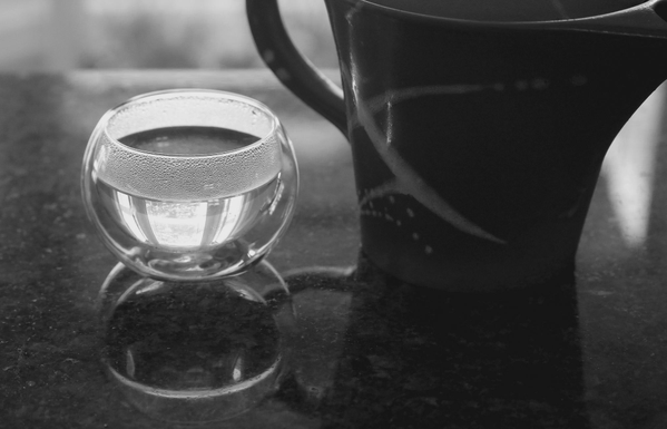 Close up photograph in black and white of a small double glass cup of tea, round with no handle, filled with a clear tea and reflecting the upside down image of a window nearby. On the right is a glazed and painted clay teapot for one, with a handle. They are side by side on a counter, where their image is also reflected.