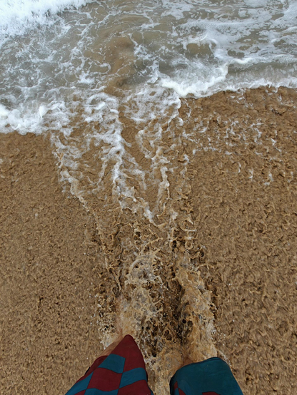 A first-person view looking down at two feet standing on the beach in the water. Receding water from the last wave is pulling and foaming around the feed, creating white foam and bubbles.