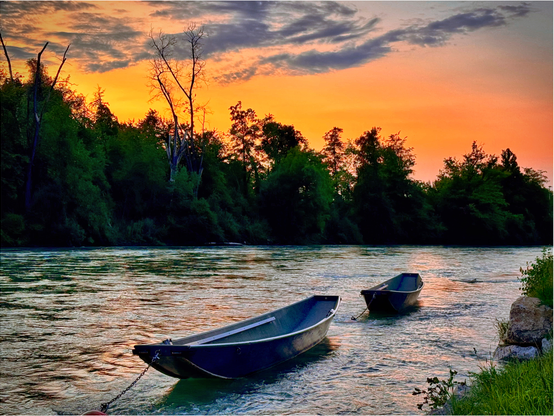 Eine friedliche Flussszene bei Sonnenuntergang mit zwei kleinen Booten, die in Ufernähe vor Anker liegen. Üppig grüne Bäume säumen das Flussufer, und der Himmel ist bei Sonnenuntergang in Orange- und Violetttönen gehalten.