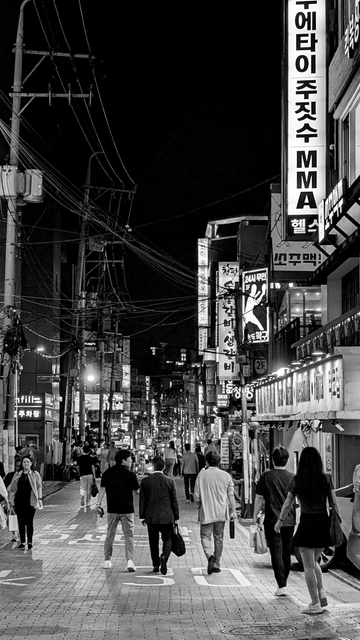 Black and white photo of a street in Gangnam, Seoul, South Korea. Image shows people walking around on a buy night among the bright lit neon lights of bars and restaurants. To left are the the crazy electrical wiring system seen all over Seoul.