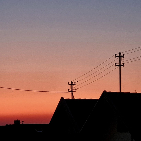 Low on the horizon, the sky is red with a touch of orange, above it a shimmering reddish blue. Two houses with pointed roofs standing side by side are visible as silhouettes  with power lines criss-crossing the sky above them.