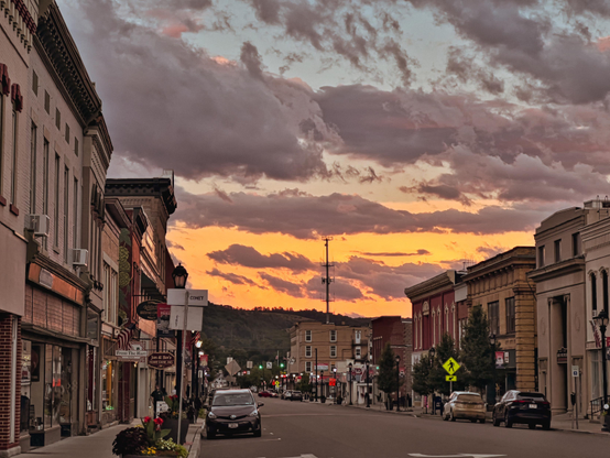 A dramatic golden hour scene captures a small-town main street with vintage brick buildings framing either side. The sky blazes with amber and purple clouds stretching across the horizon, creating a fiery canopy above the quiet street. Warm yellow light illuminates storefronts, while a few cars are parked along the weathered asphalt road that leads toward rolling hills silhouetted against the sunset.