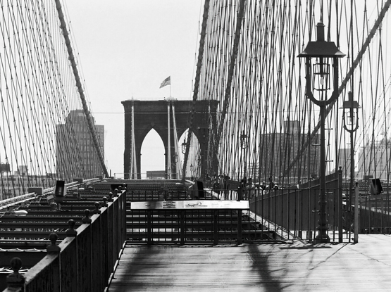 Black and white photo of the Brooklyn Bridge walkway, framed by suspension cables and arches. An American flag waves atop a distant tower.