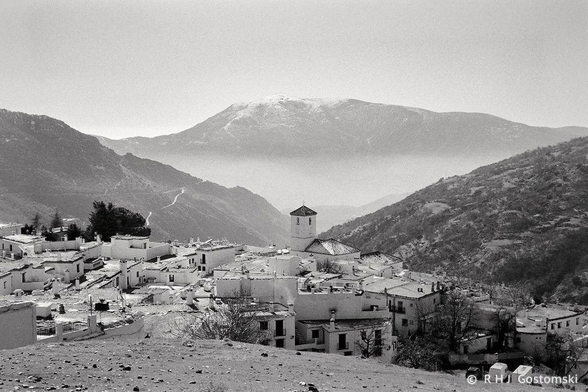A black and white photo of Capileira nestling in the Poquiera valley. A mist hangs in the valley in the distance, with mountains in the background