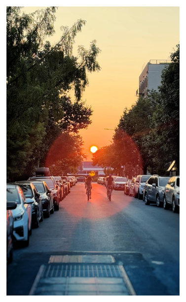 Sunset on a quiet residential street in Brooklyn, with the camera pointed straight down the street and into the sun low on the horizon. Two cyclists ride their bikes down the middle of the street towards the viewer. Parked cars and tall trees line the curbs, with the top of an apartment building poking out over the trees at right. It’s a slightly smoky day because of wildfires from Canada, turning the sky a dull yellow and giving the sun a reddish cast. A lens flare from the sun appears as a large red-orange circle in the center of the photo, enveloping the cyclists and giving everything inside its area a slightly washed-out, overexposed look.