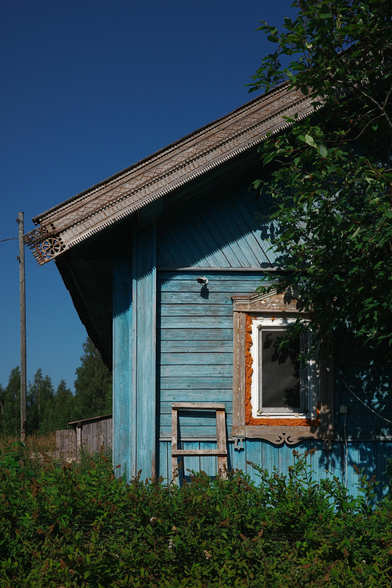 The photo shows an old wooden house with a blue color, surrounded by greenery. The carved decorations on the facade give it a special charm. However, the modern plastic window stands out, contrasting with the old appearance of the building. This element creates an interesting dissonance between the past and the present.