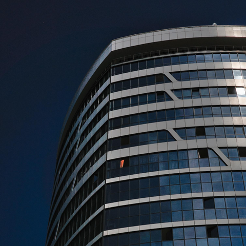 The photo shows a modern high-rise building with a glass and metal facade. The building has a rounded shape and consists of several floors. The windows reflect the sky and have a blue tint. One of the windows has a red curtain.