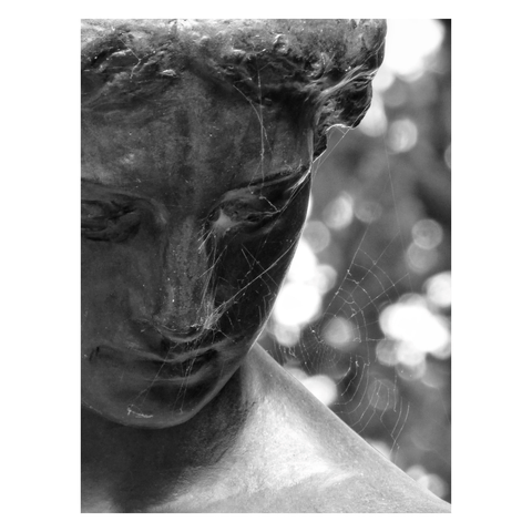 This is a black and white portrait format close-up image of a sculpture of a woman looking down. She sits to the left of the frame which cuts through her left eye. A cobweb is attached to her nose from the right, adding a sense of decay to the already melancholy feel of the image. Taken in Berlin, Germany.