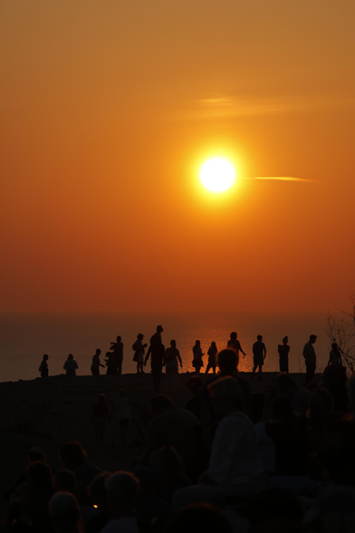 Die Sonne strahlt in der Mitte des Bildes stark gelb-orange und bestimmt durch die Überbelichtung das Bild, das fast nur orange zeigt. Im Vordergrund sind eine Reihe von Menschen nur als schwarze Schemen zu sehen. Sie laufen hintereinander auf einem Dünenkamm parallel zur Sonne. 