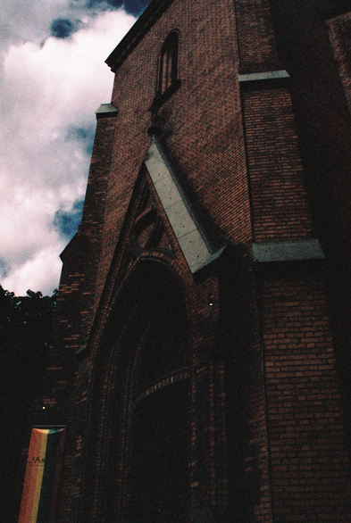 A dark and gritty photograph of the entrance to a tall and brown brick church. The brickwork has several arches and angles and is topped with copper roofing. In front of the church hangs a rainbow banner welcoming visitors. Due to the film stock's strange colour representation, the green and blue in the rainbow look almost black.