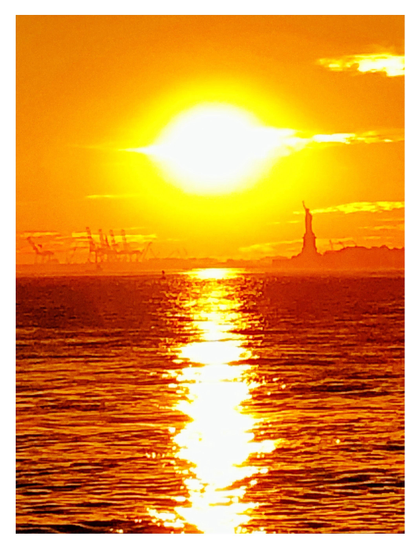 An overexposed photo of the sun setting over the East River in New York, flanked by a row of shipping cranes on the horizon to its left and the Statue of Liberty to its right. Some illuminated clouds float in from right frame. Everything is in warm bright tones of yellow, gold, and orange. The sun casts a long column of glittering light on the water, straight down the vertical center of the image.