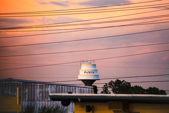 The Publix cake water tower in Lakeland, FL sits in the skyline, behind telephone poll wires. A vivid orange and pink sunset surrounds the tower.