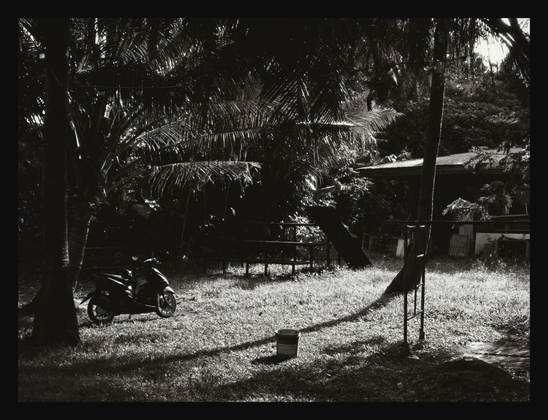 A high-contrast, black-and-white photograph captures a sunlit clearing in a tropical setting. A motorcycle is parked in the shade of dense palm trees on the left, while a lone bucket sits on the grassy ground, casting a long shadow. There's a house partially visible in the background.
