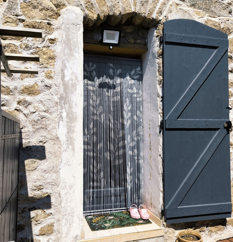 Une porte-fenêtre protégée par un rideau en bambous colorés de feuillages clairs sur un fond gris. À droite, un volet gris foncé est ouvert. À gauche, on devine une petite porte de là même couleur. Devant la porte, un paillasson coloré et une paire de Crocs roses. Le soleil tape sur le mur en pierre de la maison.