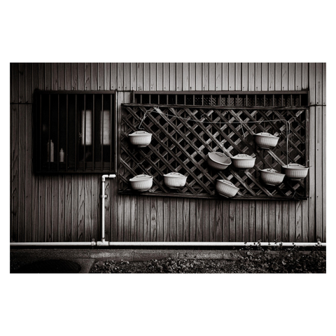 A black-and-white photograph of a wooden building wall in Nagoya. On the left, a barred window reveals faintly lit bottles inside. To the right, a lattice panel is mounted to the wall, holding several white plastic hanging pots — all empty. The trellis is perfectly aligned, the hanging wires taut, and the background wood panels run vertically with precise joins. The lack of plants or soil gives the scene a quiet, almost comic sense of futility, as though the pots are waiting for something that may never come.